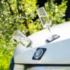 Zwei leere Sektgläser auf schwarzen quadratischen Untersetzern schräg auf der weißen Motorhaube eines Autos, unscharfer grüner Hintergrund