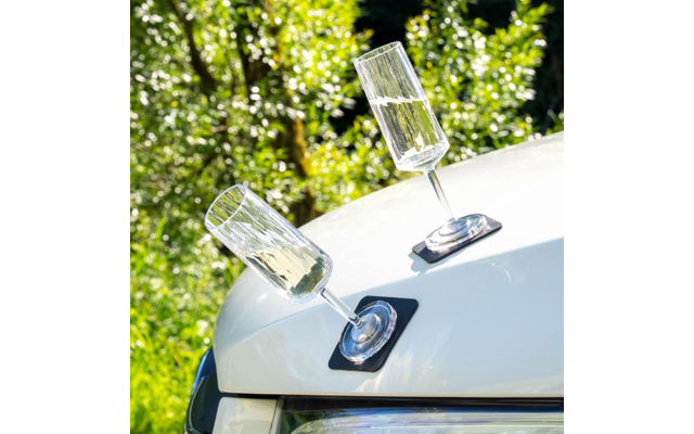 Zwei leere Sektgläser auf schwarzen quadratischen Untersetzern schräg auf der weißen Motorhaube eines Autos, unscharfer grüner Hintergrund