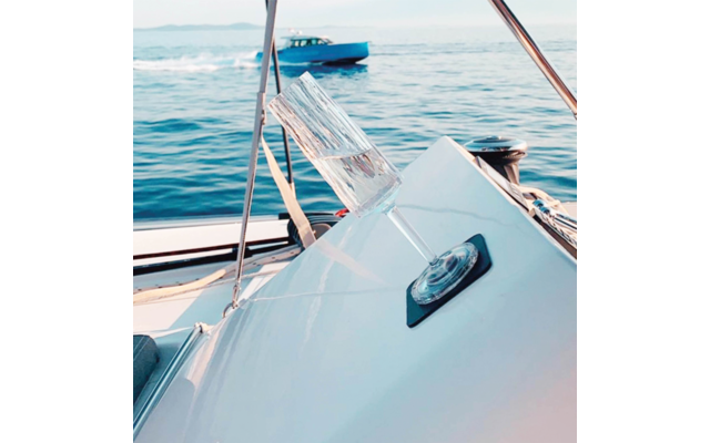 Schräg liegendes Sektglas auf einem schwarzen Untersetzer auf weißer Bootskante, Meer und Motorboot im Hintergrund