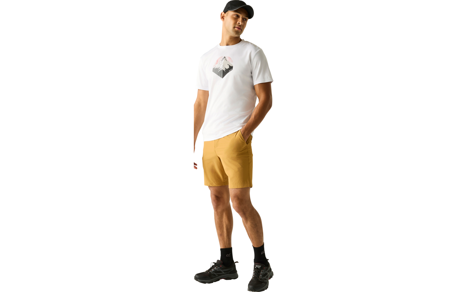 Stehender Mann mit schwarzer Cap, weißem T‑Shirt mit Bergmotiv, senfgelben Shorts, schwarzen Socken und schwarzen Sneakern vor hellem einfarbigem Hintergrund; eine Hand in der Hosentasche