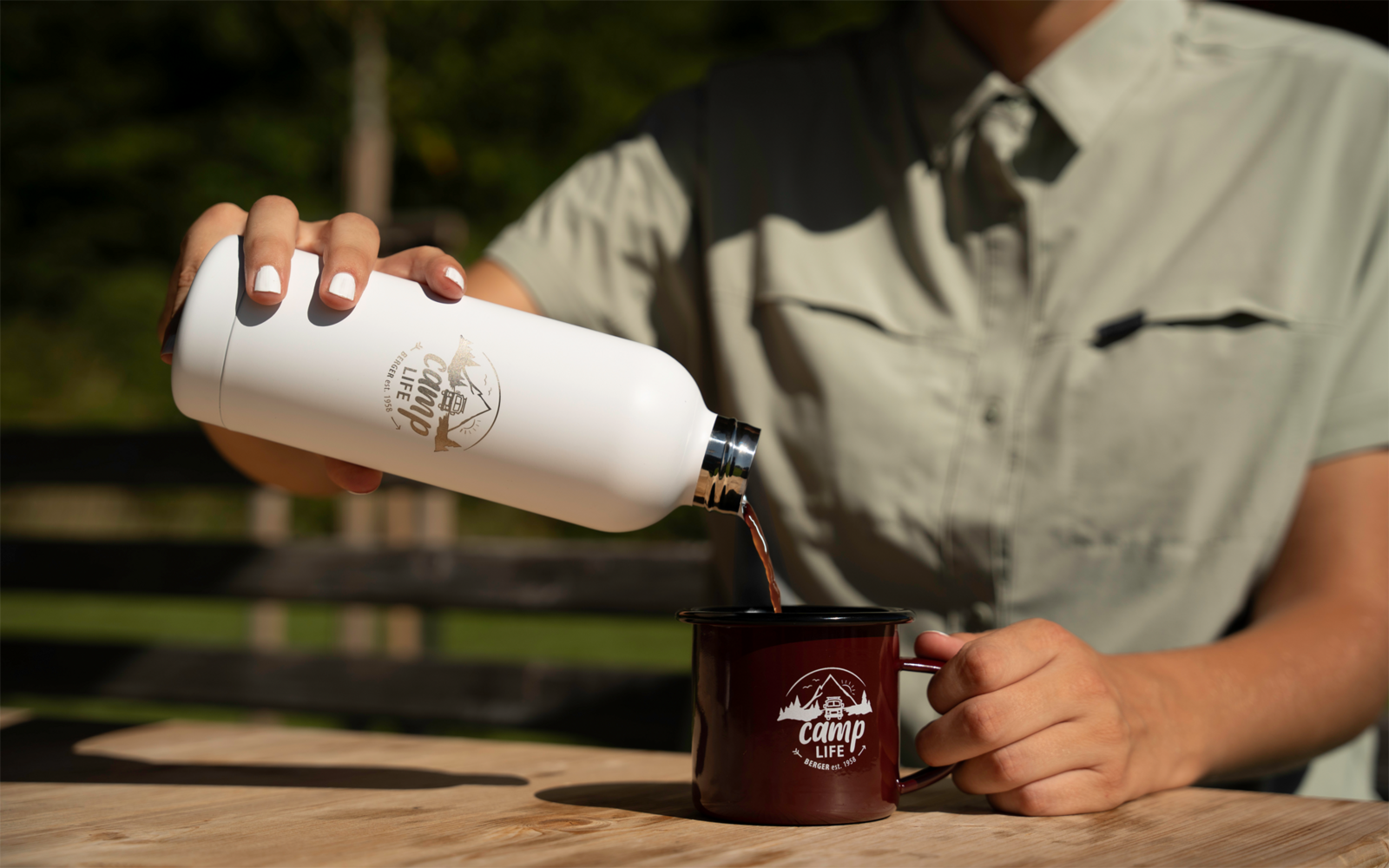 Bovenlichaam van een persoon in een kaki hemd giet donkerbruine vloeistof uit een witte thermosfles in een donkerrode emaille mok met wit logo op een houten tafel, vage tuinachtergrond