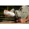 Buste d'une personne en chemise kaki versant un liquide brun foncé d'une bouteille isotherme blanche dans une tasse émaillée rouge foncé avec logo blanc sur une table en bois, arrière-plan de jardin flou