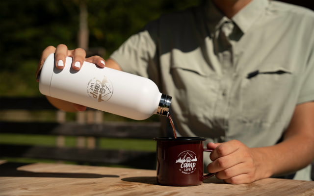 Buste d'une personne en chemise kaki versant un liquide brun foncé d'une bouteille isotherme blanche dans une tasse émaillée rouge foncé avec logo blanc sur une table en bois, arrière-plan de jardin flou
