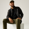 Mann sitzt auf weißem Würfel, trägt Basecap, schwarze Jacke mit Camouflageärmeln und olivgrüne Hose, blickt nach oben rechts