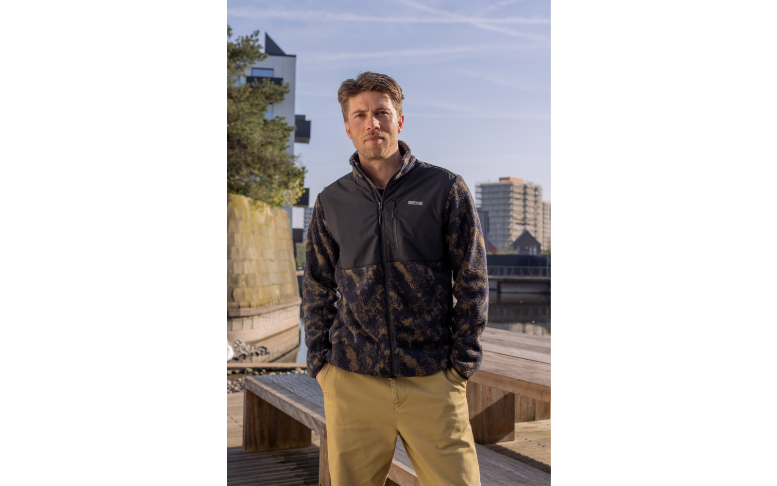 Man staat op een houten steiger bij het water, draagt een zwarte gevlekte fleecejas en een beige broek; gebouwen en blauwe lucht met condenssporen op de achtergrond
