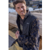 Man zit op een houten steiger aan het water, kijkt naar links en lacht; hij draagt een gemêleerd fleecejack met zwart voorpand en kaki broek, boten op de achtergrond