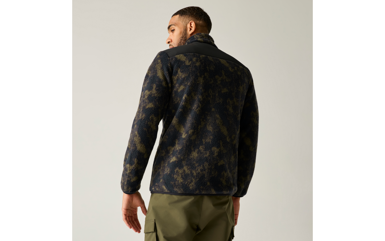 Rückansicht eines Mannes mit kurzem Haar und Bart, er trägt eine dunkle, camouflagemusterte Fleecejacke mit schwarzem Schulterbesatz und olivgrünen Cargohosen vor hellem Hintergrund