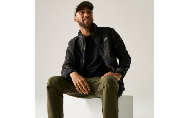 Mann sitzt auf weißem Würfel, trägt Basecap, schwarze Jacke mit Camouflageärmeln und olivgrüne Hose, blickt nach oben rechts