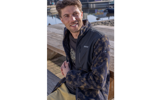 Man zit op een houten steiger aan het water, kijkt naar links en lacht; hij draagt een gemêleerd fleecejack met zwart voorpand en kaki broek, boten op de achtergrond
