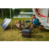 Mann sitzt vor einem Wohnwagen auf einem Campingstuhl, ein Tisch mit Essen und Getränken steht davor, daneben sind ein Solarpanel und eine Kühlbox