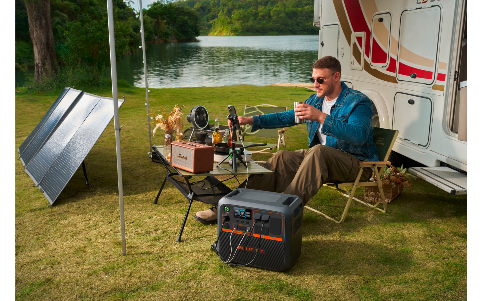 Mann sitzt vor einem Wohnwagen auf einem Campingstuhl, ein Tisch mit Essen und Getränken steht davor, daneben sind ein Solarpanel und eine Kühlbox