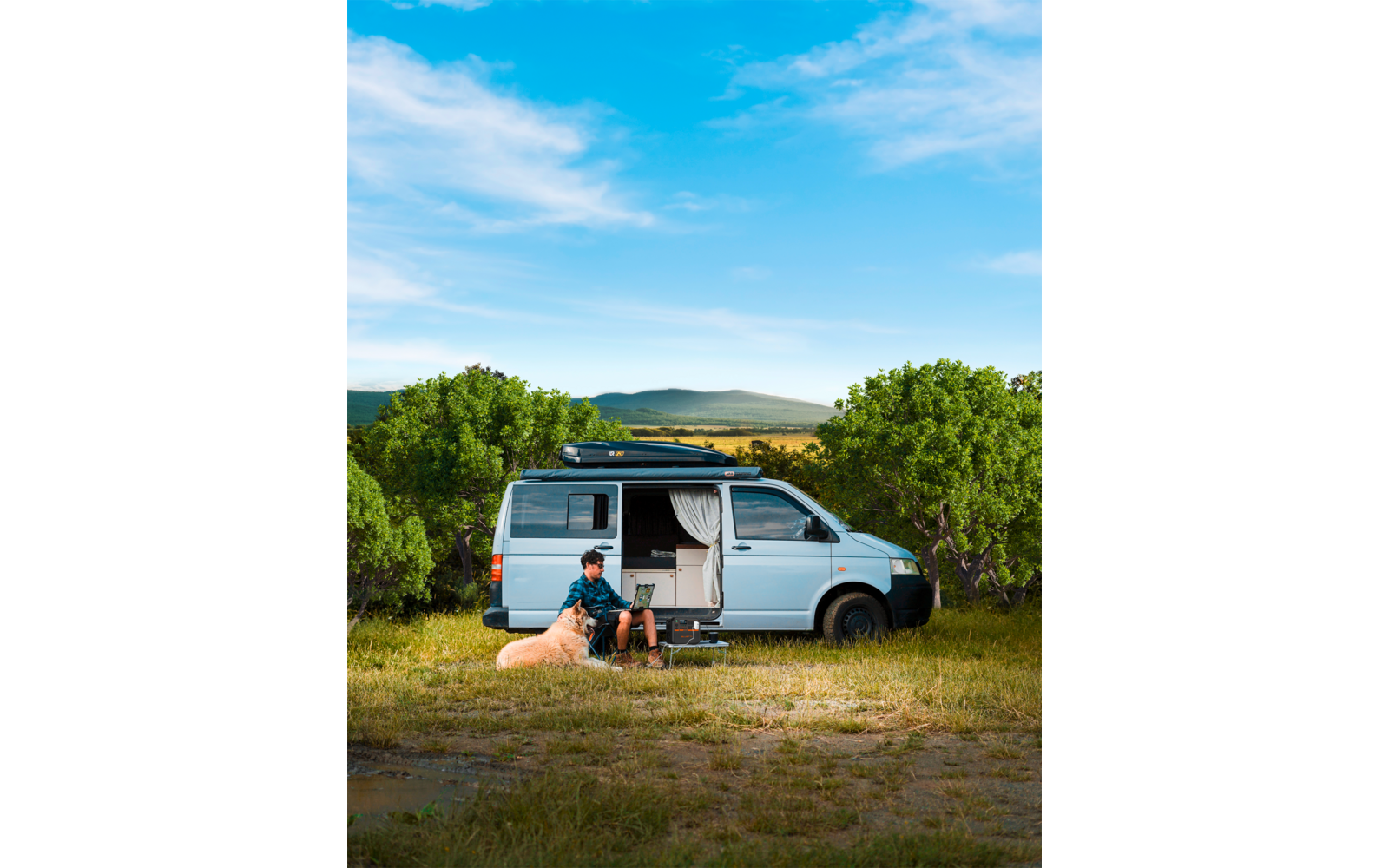 Ein Wohnmobil steht auf einer Wiese, daneben sitzt eine Person auf einem Stuhl unter blauem Himmel