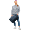 Frau steht, trägt hell gestreiftes Langarmshirt, blaue Jeans, weiße Sneaker, Sonnenbrille auf dem Kopf, hält dunkelblaue Reisetasche mit Logo deuter, Haare zu Pferdeschwanz, lächelnd