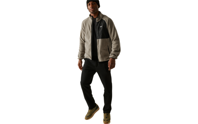 Mann trägt eine Mütze, eine zweifarbige Jacke, schwarze Hose und braune Schuhe, steht vor weißem Hintergrund