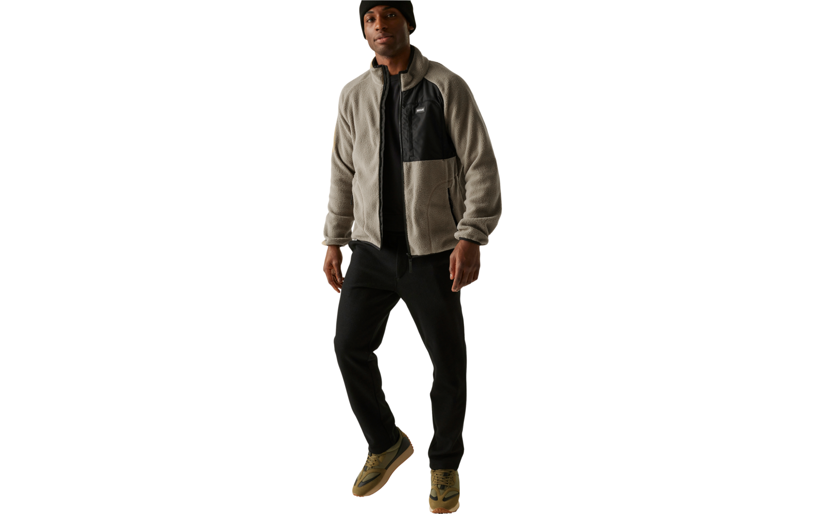 Mann trägt eine Mütze, eine zweifarbige Jacke, schwarze Hose und braune Schuhe, steht vor weißem Hintergrund