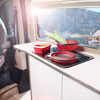 Wohnmobil-Küchenzeile mit schwarzem Kochfeld, zusammenklappbaren roten Silikon-Schüsseln und einem verschlossenen roten Behälter; grüne Frühlingszwiebeln liegen darauf; Fenster mit Bergen, See und Sonnenlicht