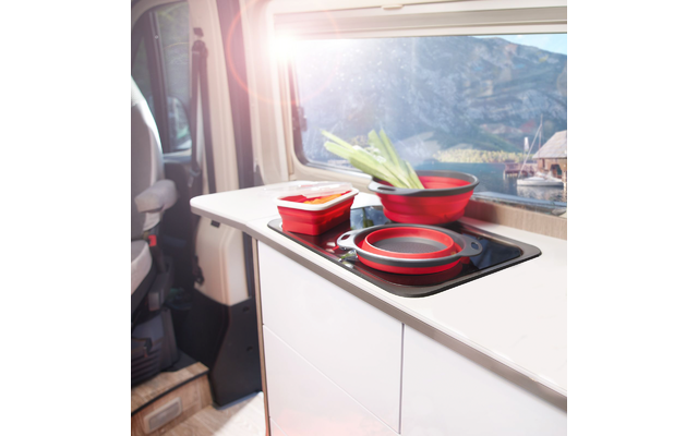 Wohnmobil-Küchenzeile mit schwarzem Kochfeld, zusammenklappbaren roten Silikon-Schüsseln und einem verschlossenen roten Behälter; grüne Frühlingszwiebeln liegen darauf; Fenster mit Bergen, See und Sonnenlicht