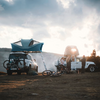 Deux VUS avec tentes de toit déployées, vélos fixés à un véhicule, matériel de camping et des personnes sur un terrain découvert, collines et ciel nuageux en arrière-plan