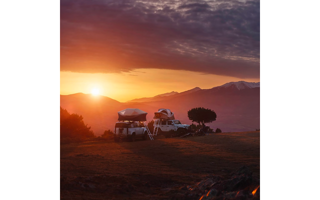 Deux SUV avec tentes de toit et échelles sur un terrain herbeux devant des montagnes, un arbre isolé à côté des véhicules, soleil près de l'horizon, ciel nuageux