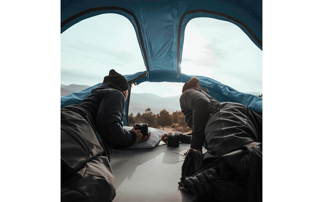 Deux personnes allongées dans une tente, visibles de dos, regardant par l'ouverture de la tente un paysage montagneux ; deux tasses et un appareil photo à côté d'eux