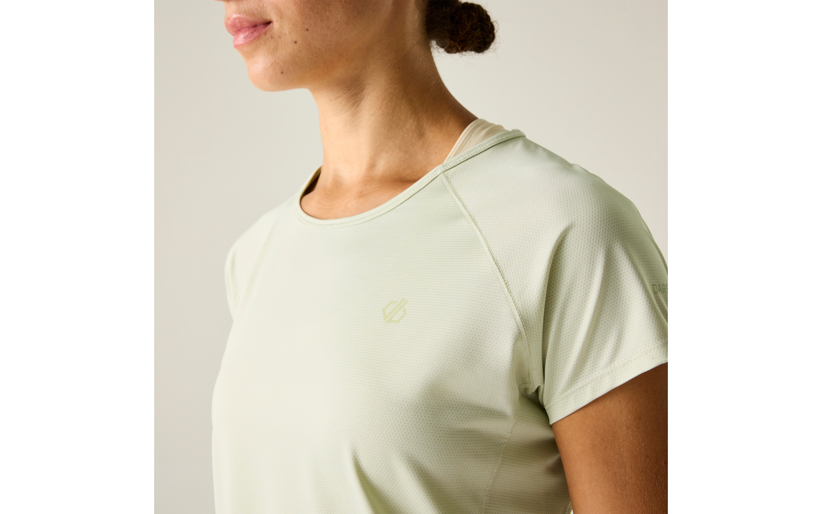 Nahaufnahme des Oberkörpers und des unteren Gesichts einer Person mit hellgrünem, kurzärmeligem Sportshirt und kleinem Logo auf der Brust; Haare zu einem niedrigen Dutt; heller einfarbiger Hintergrund