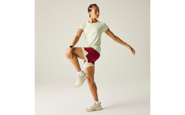 Frau steht auf einem Bein, rechtes Knie angezogen, rechte Hand am Knie, linker Arm seitlich ausgestreckt; trägt hellgrünes T‑Shirt, dunkelrote Shorts mit hellen Einsätzen, beige Sneaker und Smartwatch; neutraler Hintergrund