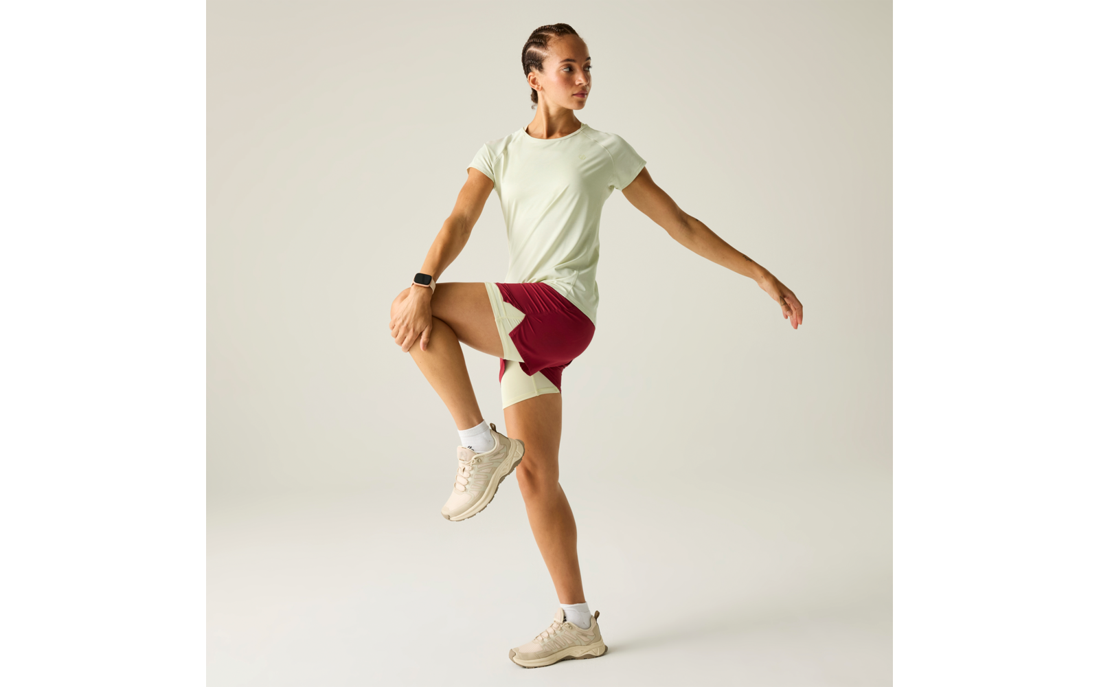 Frau steht auf einem Bein, rechtes Knie angezogen, rechte Hand am Knie, linker Arm seitlich ausgestreckt; trägt hellgrünes T‑Shirt, dunkelrote Shorts mit hellen Einsätzen, beige Sneaker und Smartwatch; neutraler Hintergrund