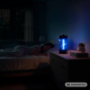 Persona addormentata nel letto; comodino con un dispositivo che emette luce blu, pianta in vaso e libri; finestra con tende sullo sfondo
