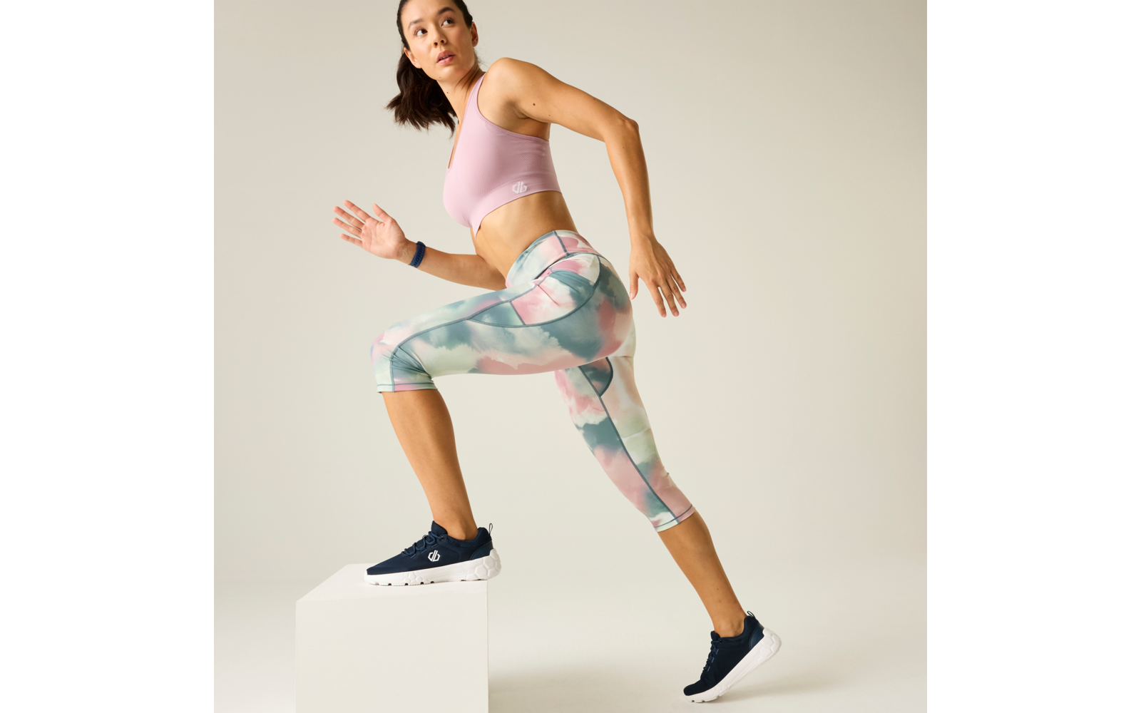 Person in a pink sports bra and pastel patterned capri leggings stepping with one foot onto a white block; navy sneakers, hair tied back, plain light background
