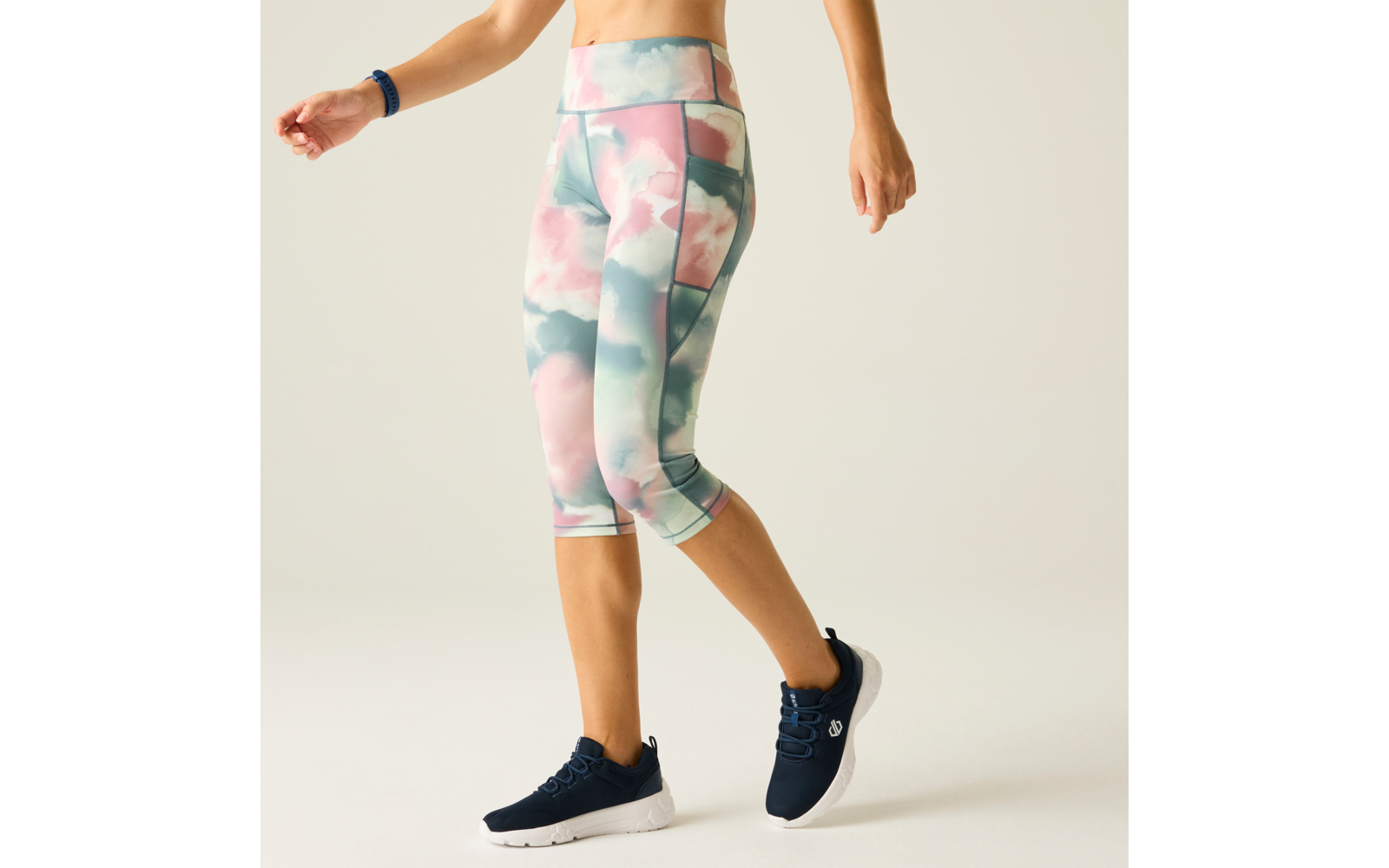Lower torso and legs of a person wearing pastel floral capri leggings and navy sports shoes; right wrist with a fitness band; light background