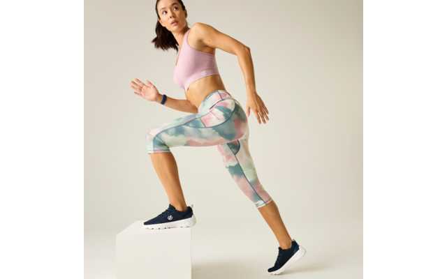 Person in a pink sports bra and pastel patterned capri leggings stepping with one foot onto a white block; navy sneakers, hair tied back, plain light background