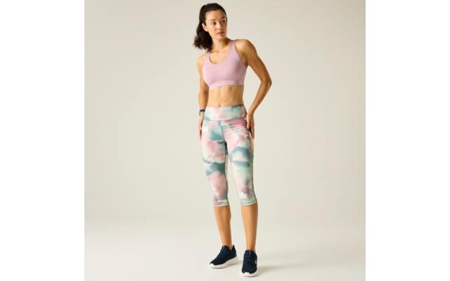 Woman wearing a pink sports bra and pastel patterned capri leggings, standing with one hand on her hip and looking to the right against a light background