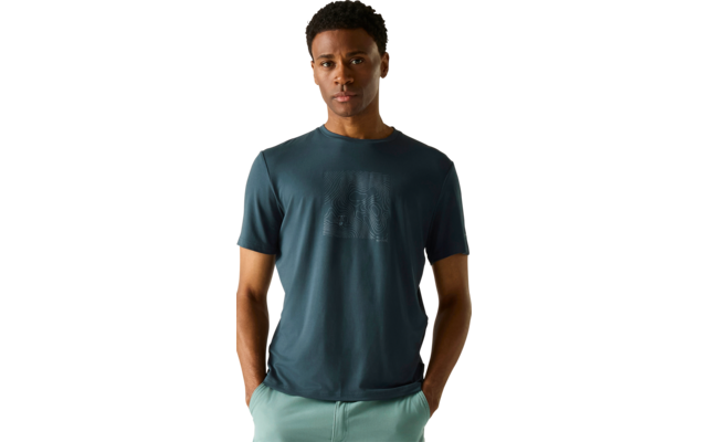 stehende Person, frontal zur Kamera, trägt ein dunkel-türkisfarbenes Kurzarmshirt mit geprägtem Muster auf der Brust, helle grüne Hose, Hände in den Taschen, weißer Hintergrund