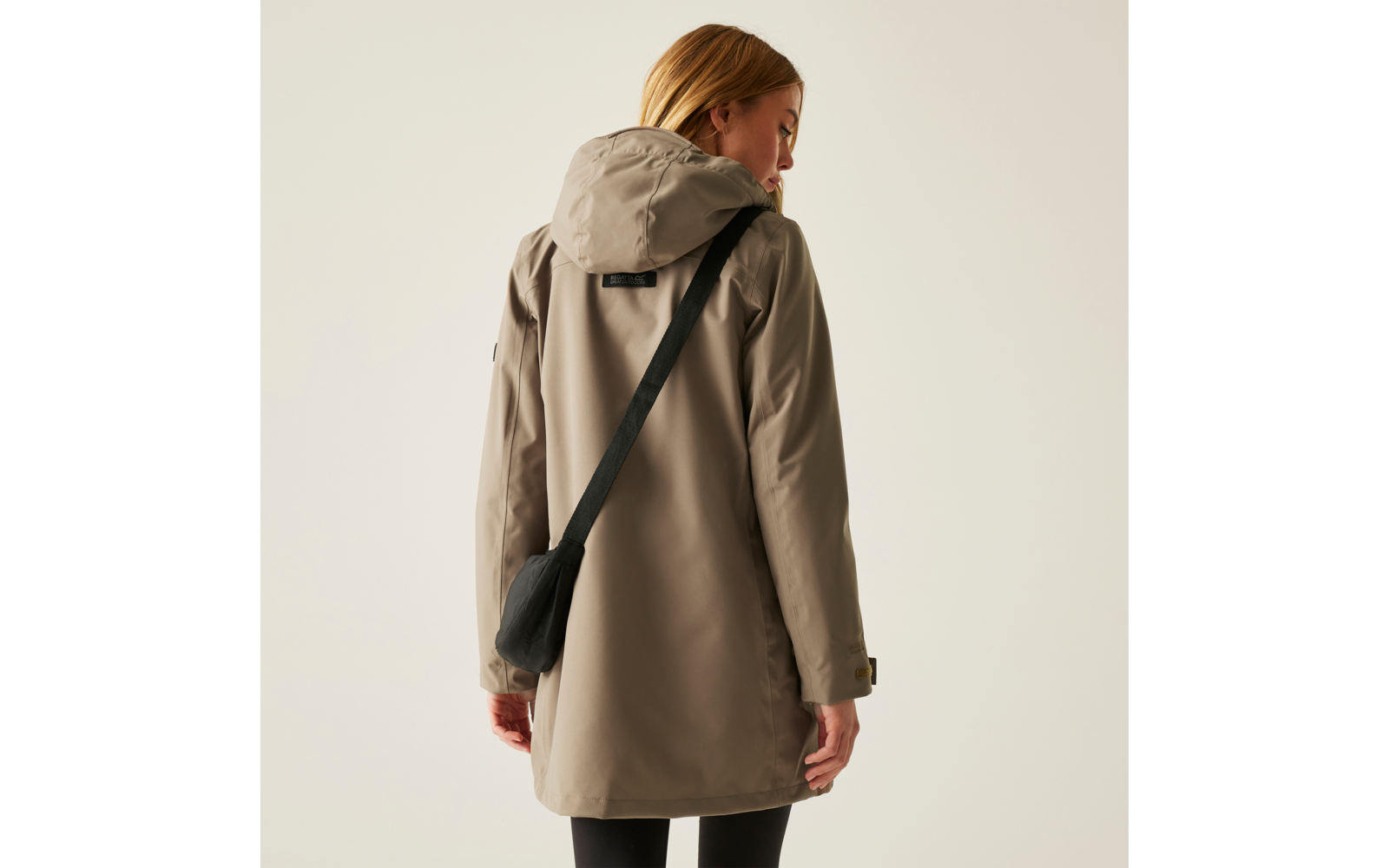 Frau mit langem blonden Haar trägt eine beige Jacke mit Kapuze und eine schwarze Umhängetasche, von hinten fotografiert