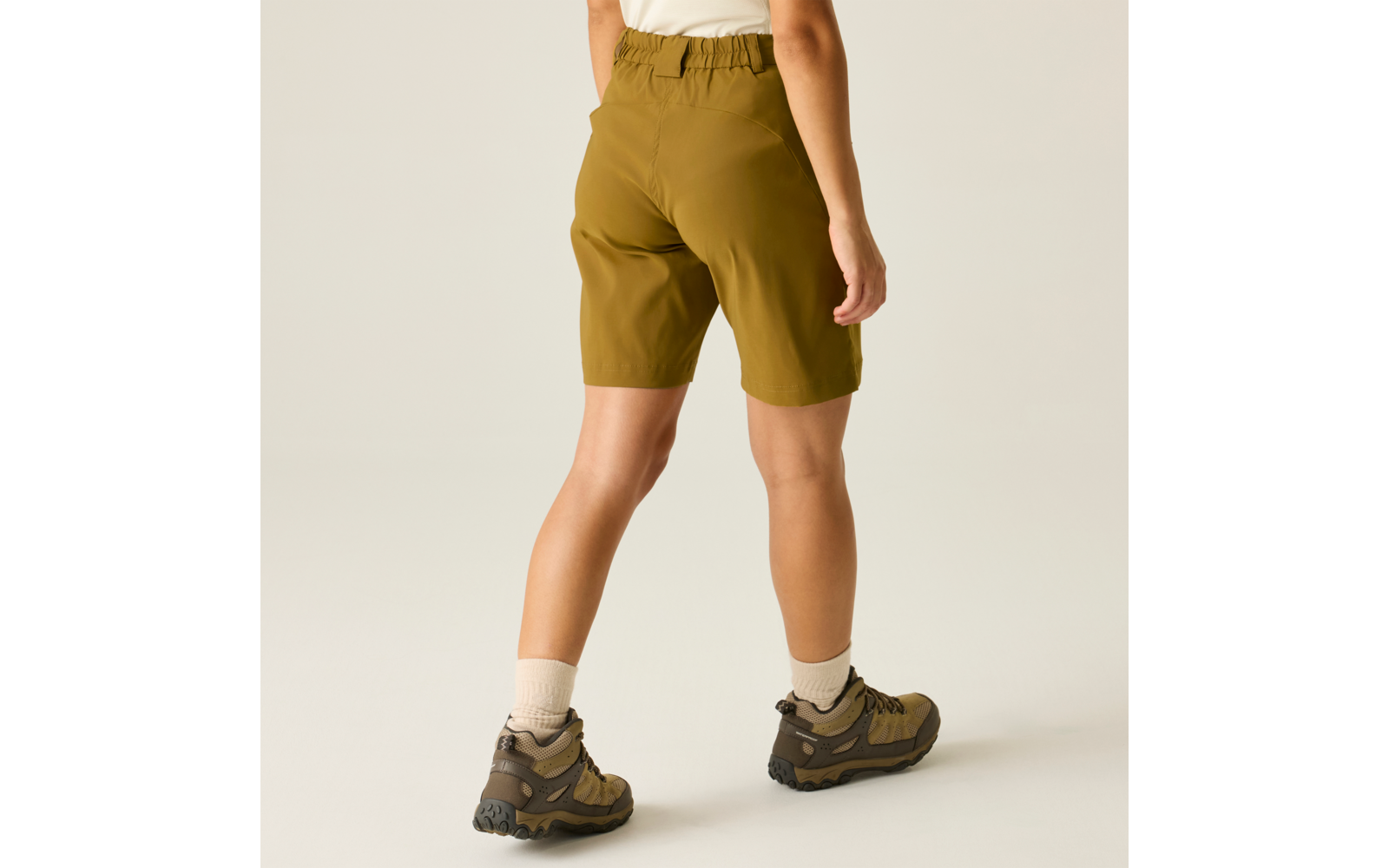 Persona vista de espaldas con pantalones cortos oliva hasta la rodilla, camiseta blanca, calcetines blancos y zapatillas de senderismo marrones sobre fondo claro