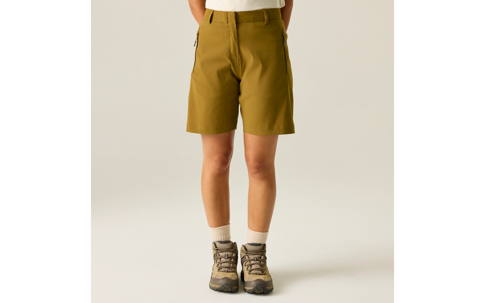 Parte inferior del cuerpo de una persona desde la cintura hasta los pies, pantalones cortos outdoor color oliva, calcetines blancos, botas de senderismo marrones, fondo claro liso