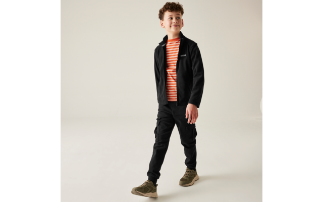 Bambino con capelli ricci che cammina verso destra, indossa giacca nera, maglietta a righe arancione e bianca, pantaloni cargo neri e sneakers verdi su sfondo chiaro