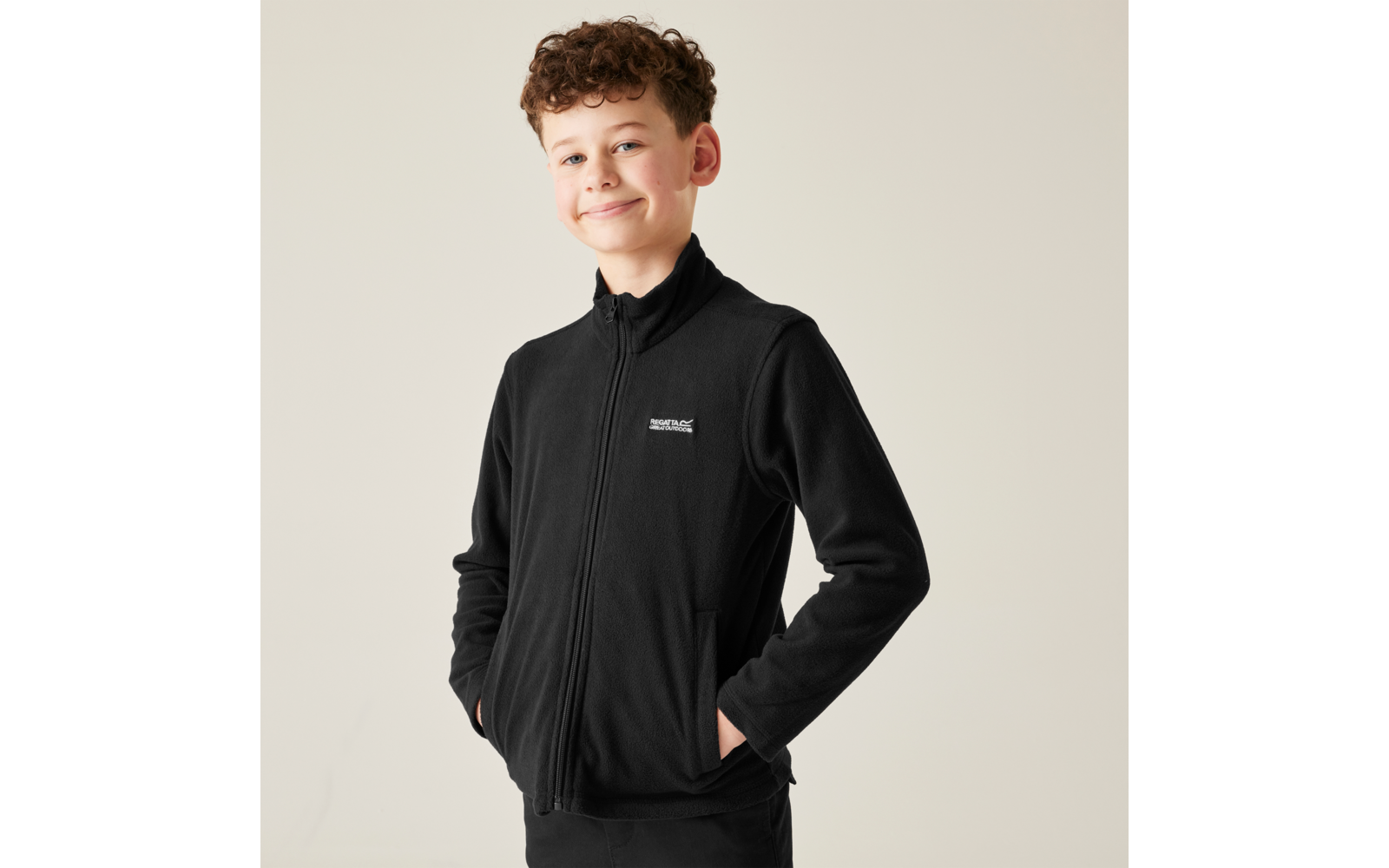 Ragazzo con capelli castani ricci che indossa una giacca in pile nera, mani nelle tasche, sfondo chiaro uniforme