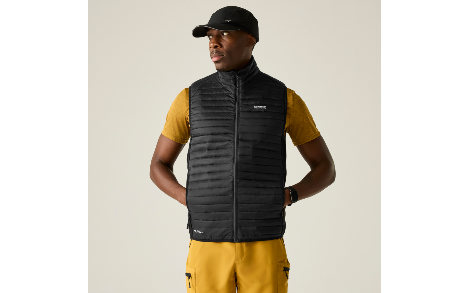Person steht vor hellem Hintergrund, trägt schwarze gesteppte Weste mit Reißverschluss, gelbes T‑Shirt, gelbe Cargohose, schwarze Kappe und Armbanduhr; Hände in den Hosentaschen