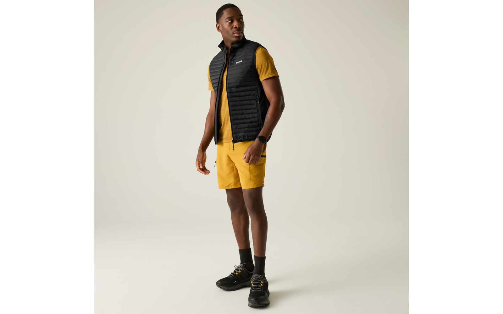 Mann steht in gelbem T-Shirt und Shorts, schwarze gesteppte Weste, schwarze Schuhe, Blick nach rechts