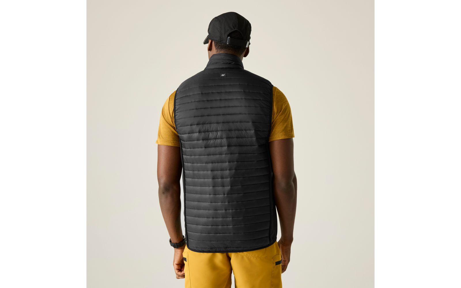 Person von hinten mit schwarzer gesteppter Weste, gelbem Kurzarmshirt, gelber Hose, schwarzer Kappe und Armbanduhr, heller einfarbiger Hintergrund