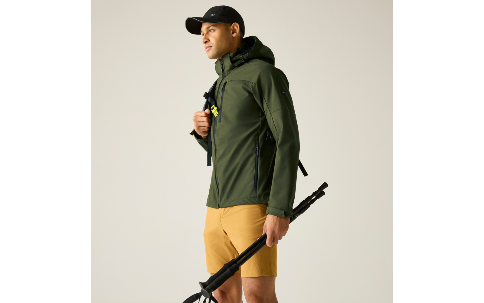 Person in olivgrüner Kapuzenjacke und schwarzer Kappe, gelbe Shorts, hält zusammengeklappte Trekkingstöcke in der rechten Hand; Rucksackriemen sichtbar; heller einfarbiger Hintergrund; schaut nach links