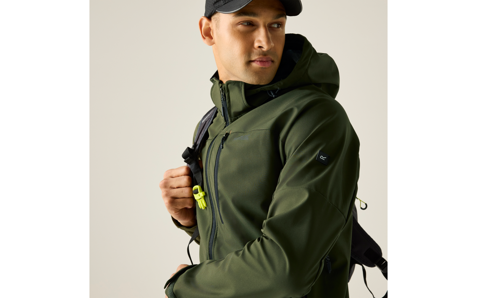 Person in olivgrüner Kapuzenjacke und schwarzer Kappe, trägt Rucksack, hält Rucksackriemen, schaut nach rechts