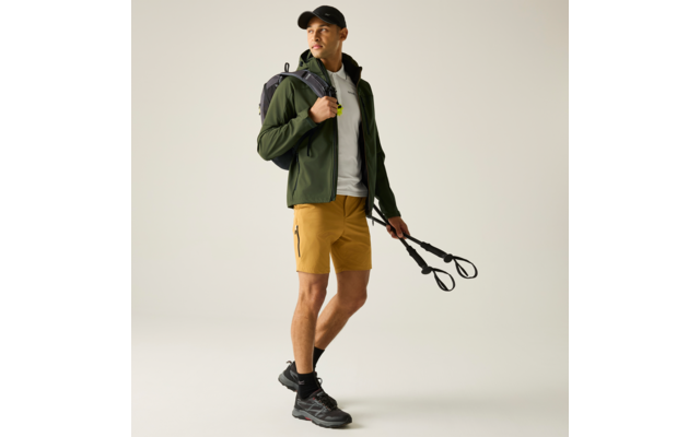 Person trägt grüne Jacke, weißes T-Shirt, senfgelbe Shorts und schwarze Cap; schwarzer Rucksack über einer Schulter; hält zwei schwarze Wanderstöcke; schaut zur Seite; heller Hintergrund