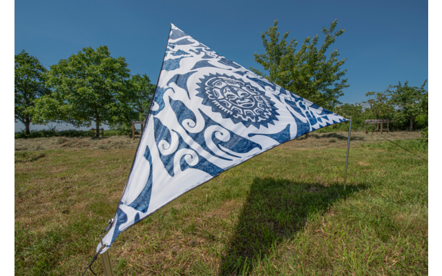 Dreieckiges Segel mit blauem Muster auf einer Wiese, im Hintergrund Bäume und blauer Himmel