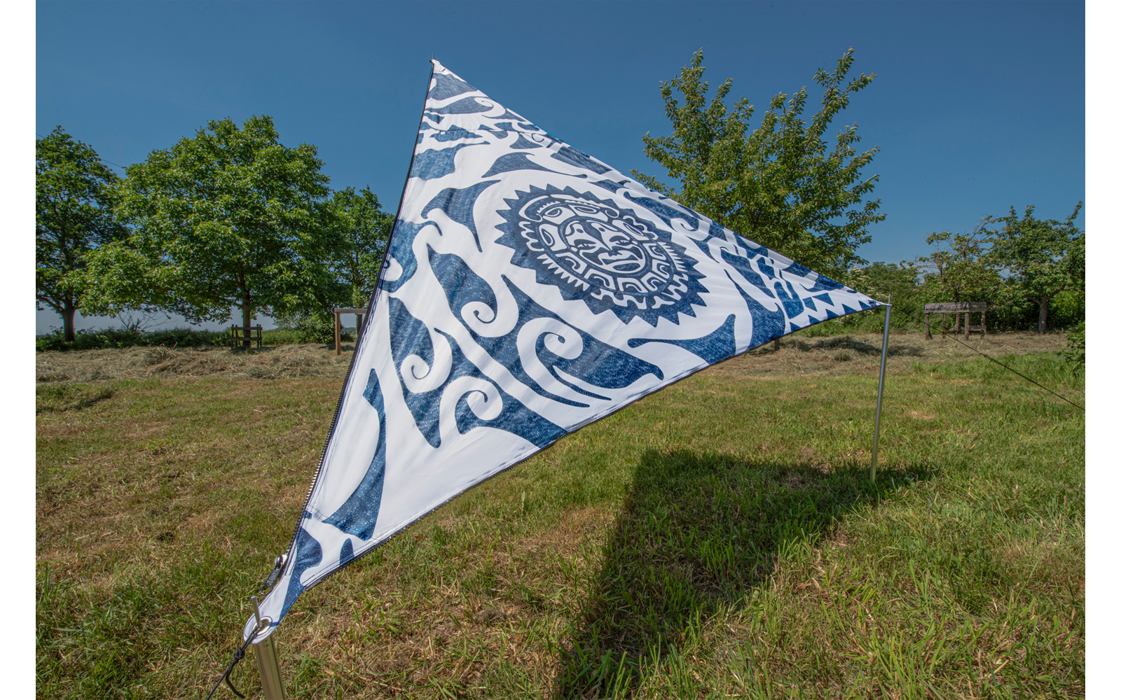 Dreieckiges Segel mit blauem Muster auf einer Wiese, im Hintergrund Bäume und blauer Himmel