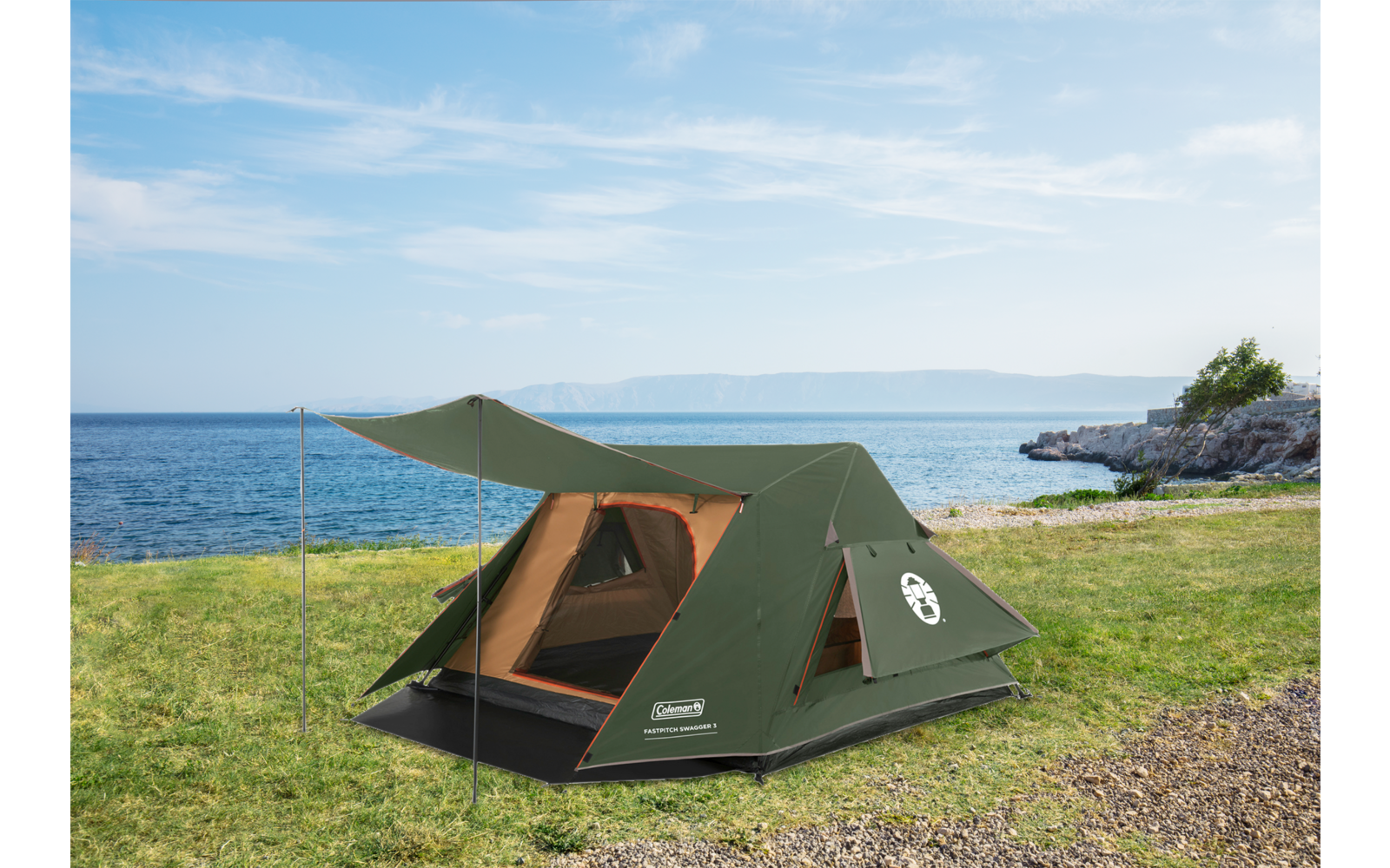 Green tent with open entrance on grass by a rocky shoreline and the sea under a partly cloudy sky