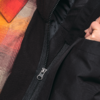 Nahaufnahme eines Reißverschlusses an einer schwarzen Jacke, darunter ein kariertes orange-rotes Hemd und eine Hand an der Jackenöffnung