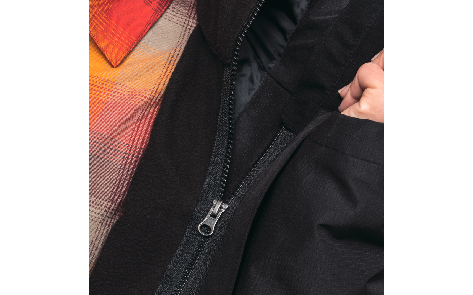 Nahaufnahme eines Reißverschlusses an einer schwarzen Jacke, darunter ein kariertes orange-rotes Hemd und eine Hand an der Jackenöffnung