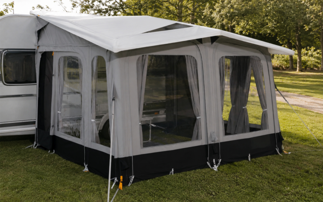 Witte caravan met grijs voortentluifel en grote gaasramen, op gras met bomen op de achtergrond